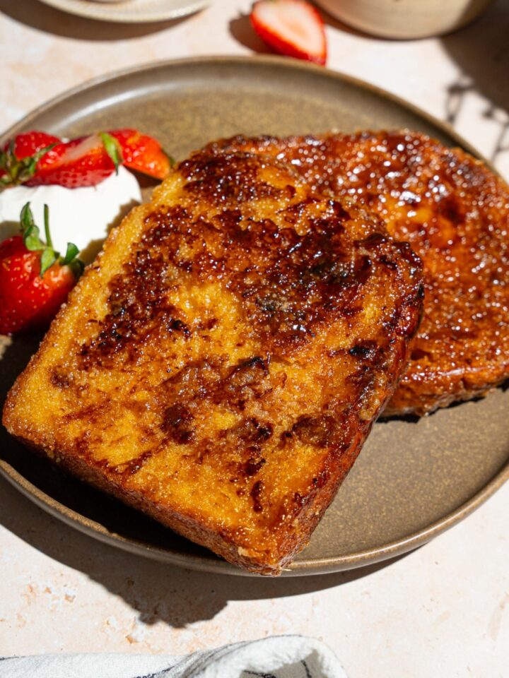 A plate with slices of creme brulee french toast served with sliced strawberries and whipped cream. The plate is on a tan counter with a bowl of strawberries.
