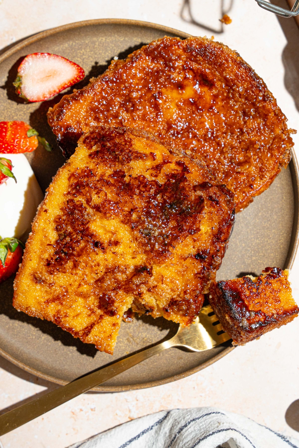 A plate with slices of creme brulee french toast served with sliced strawberries and whipped cream. A fork is taking a bite of french toast. The plate is on a tan counter.