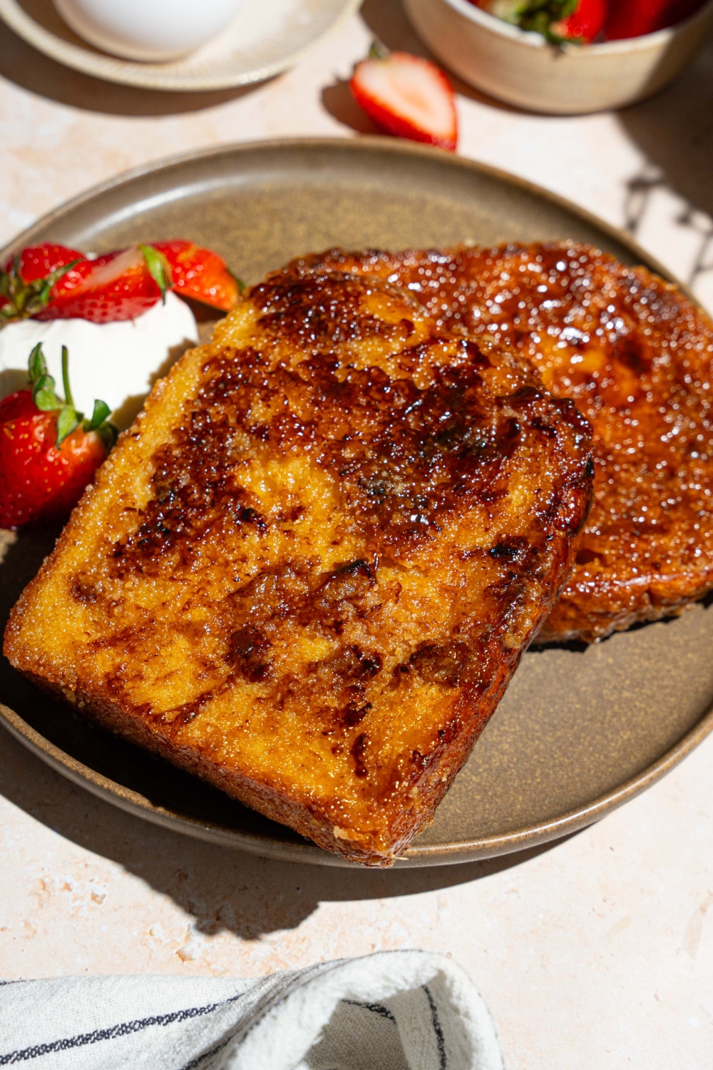 A plate with slices of creme brulee french toast served with sliced strawberries and whipped cream. The plate is on a tan counter with a bowl of strawberries.
