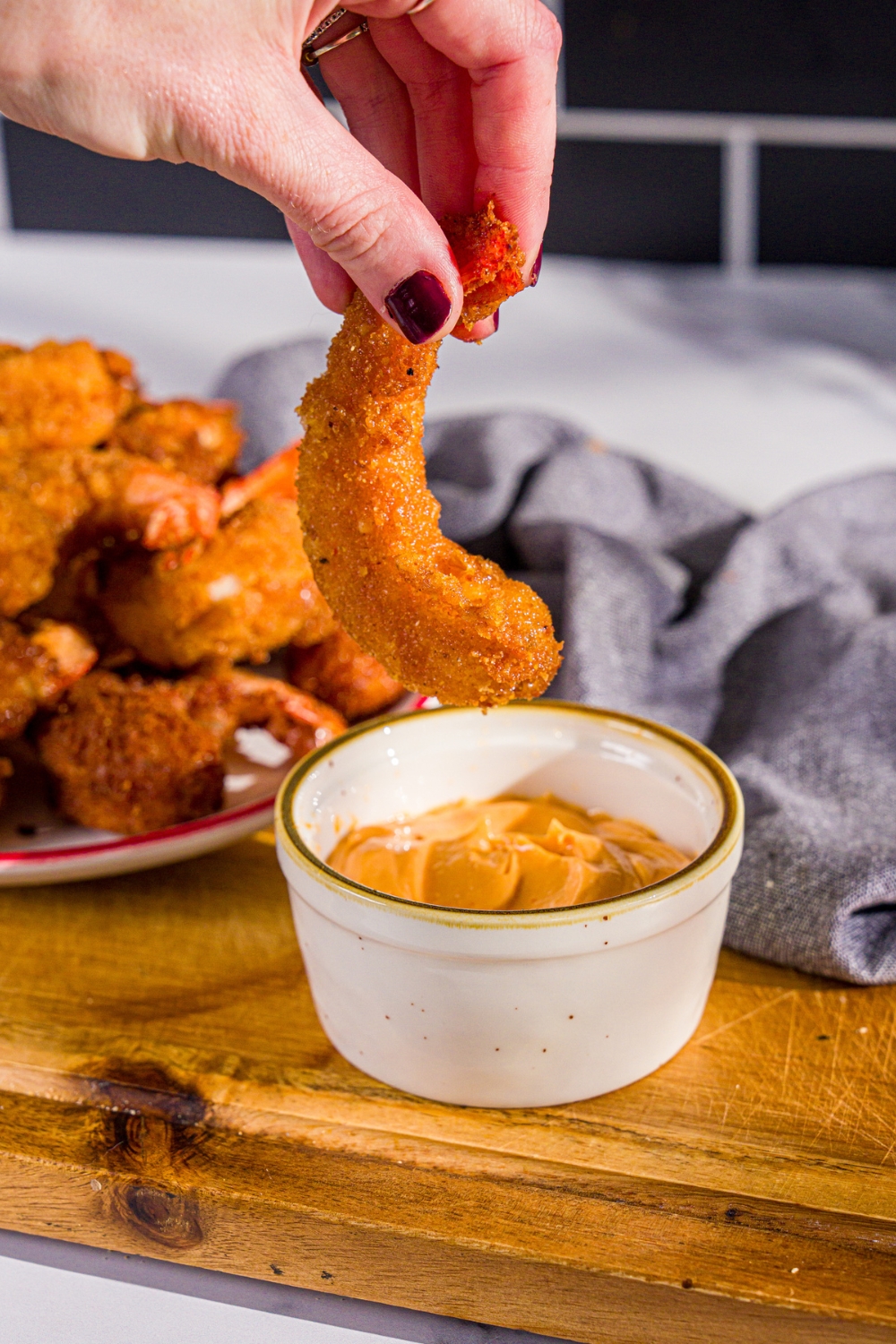 A hand holding a crispy fried shrimp above a bowl of spicy mayo that's on a wooden board on a white counter.