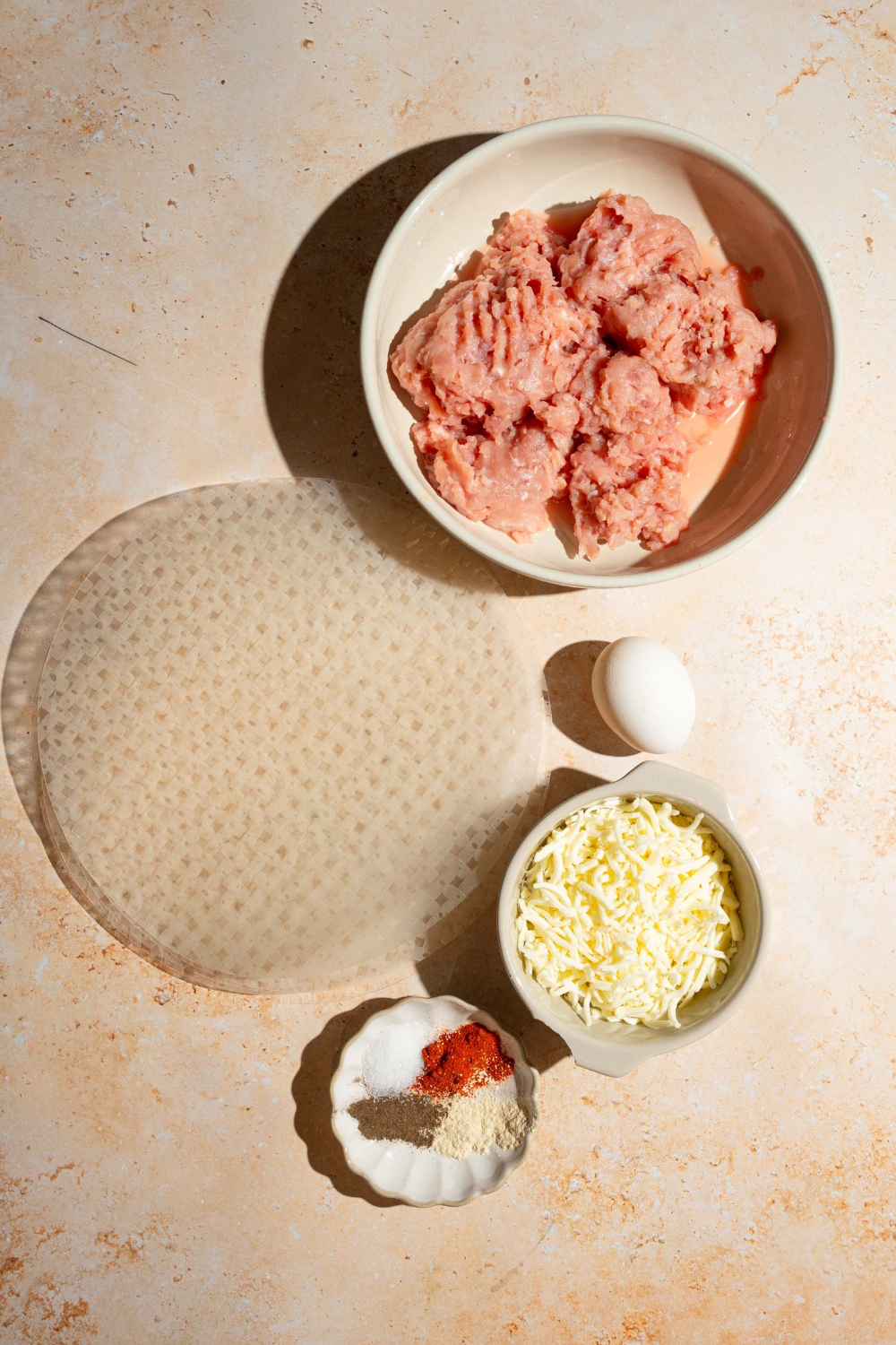 A tan counter with several bowls containing ingredients to make rice paper chicken bites including ground chicken, rice paper sheets, shredded mozzarella, and seasonings.