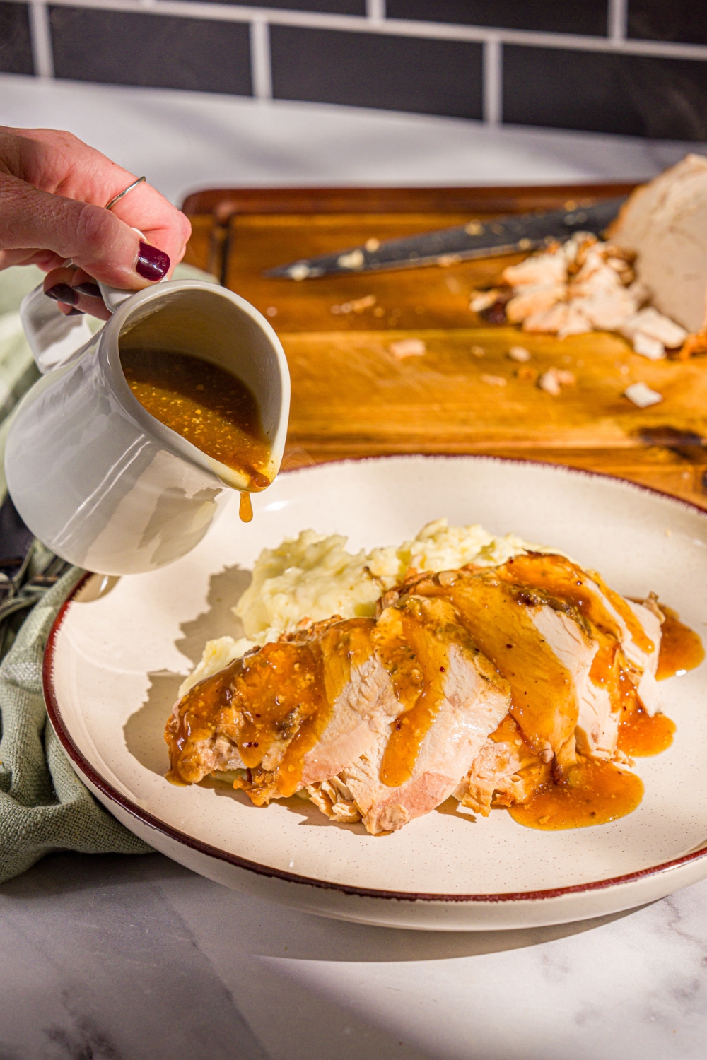 A plate of sliced crock pot butterball turkey breast with mashed potatoes. A pitcher of gravy is being poured over the turkey. The plate is on a marble counter with a wooden cutting board with turkey.