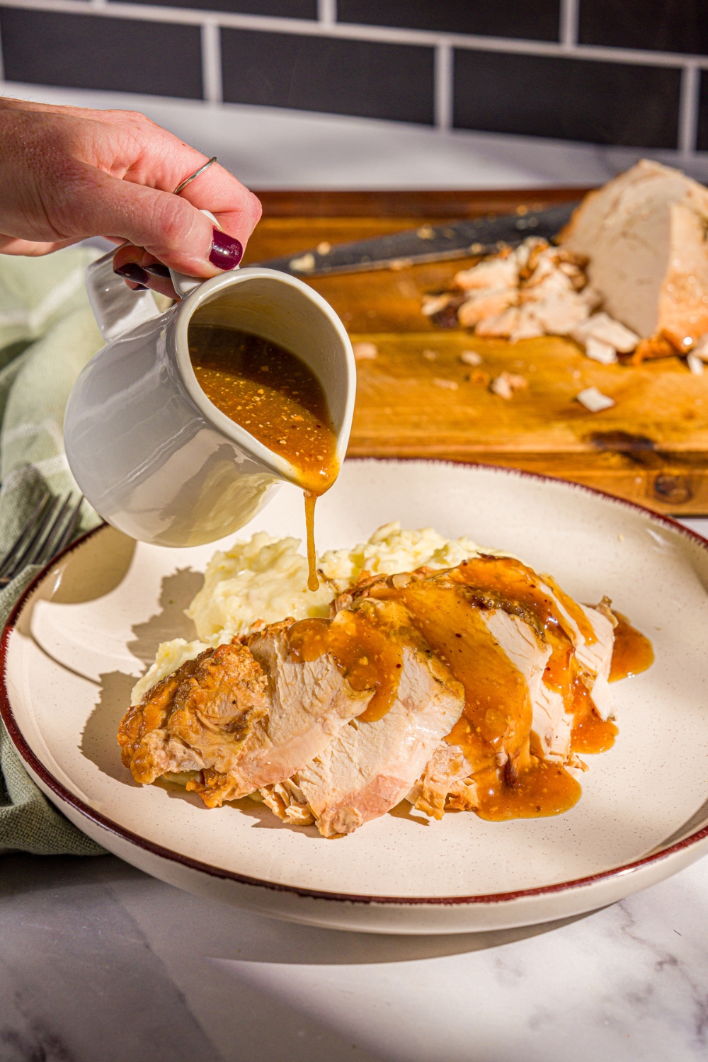 A plate of sliced crock pot butterball turkey breast with mashed potatoes. A pitcher of gravy is being poured over the turkey. The plate is on a marble counter with a wooden cutting board with turkey.
