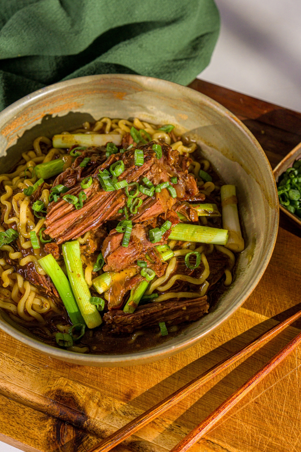 A bowl of crockpot Mongolian beef served with noodles and garnished with sliced green onion and sesame seeds. The bowl is on a wooden board with a pair of chopsticks.