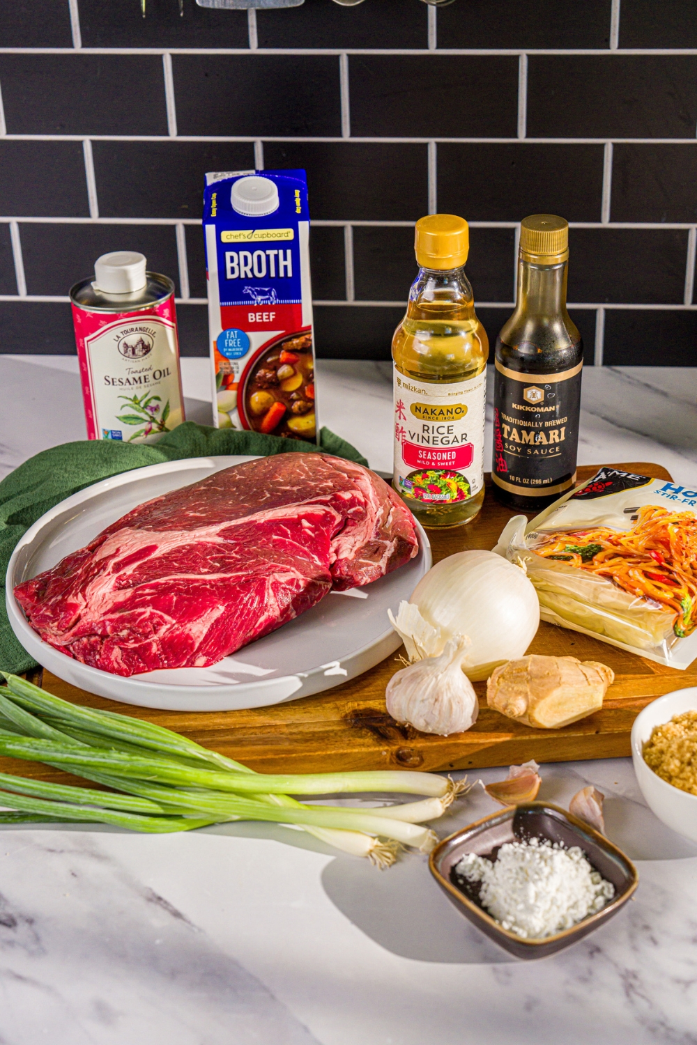 A wooden board with ingredients to make crockpot Mongolian beef including chuck roast, onion, brown sugar, soy sauce, cornstarch, beef broth, rice vinegar, garlic, ginger, tamari, and more.
