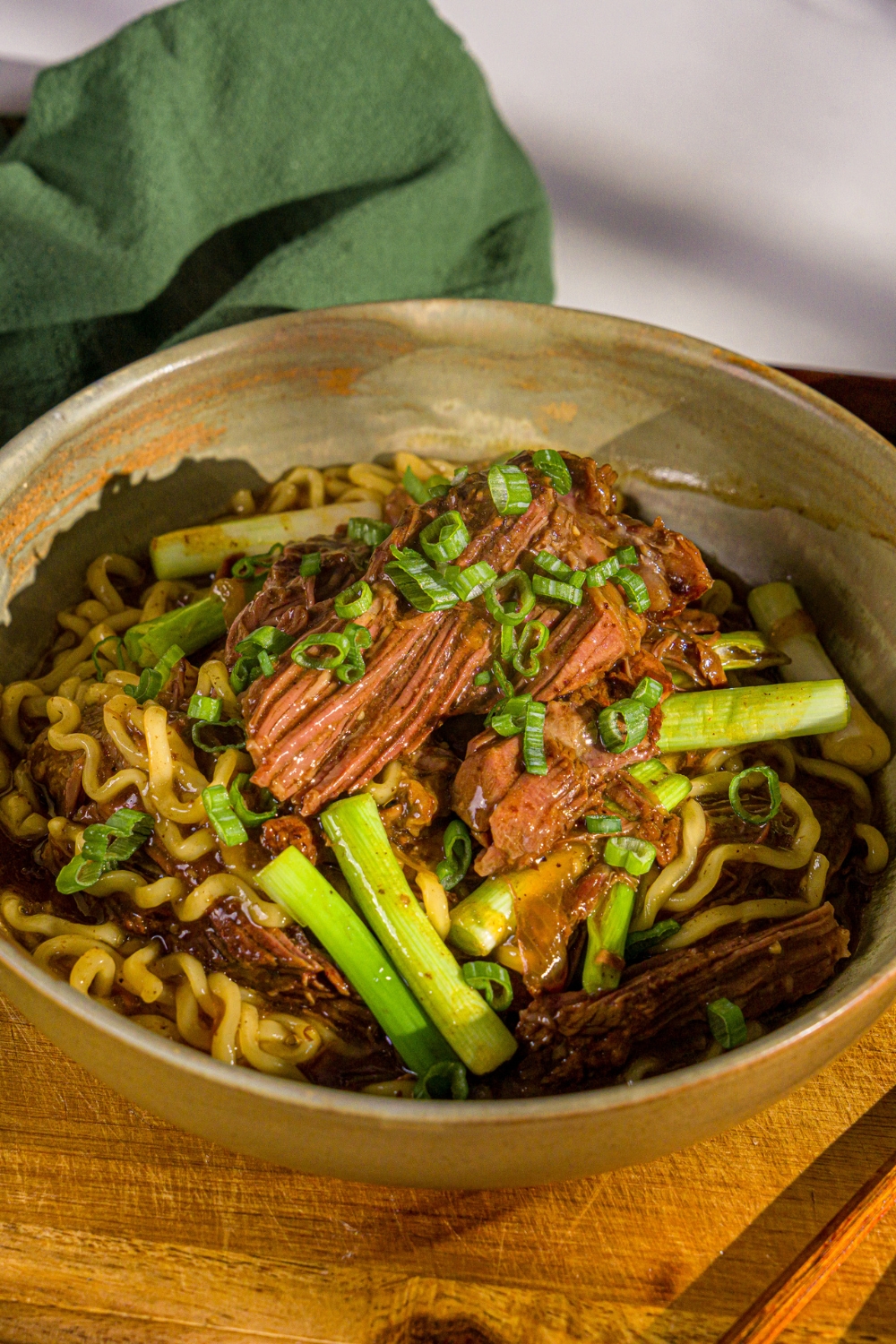 A bowl of crockpot Mongolian beef served with noodles and garnished with sliced green onions and sesame seeds. The bowl is on a wooden board with a green cloth napkin.