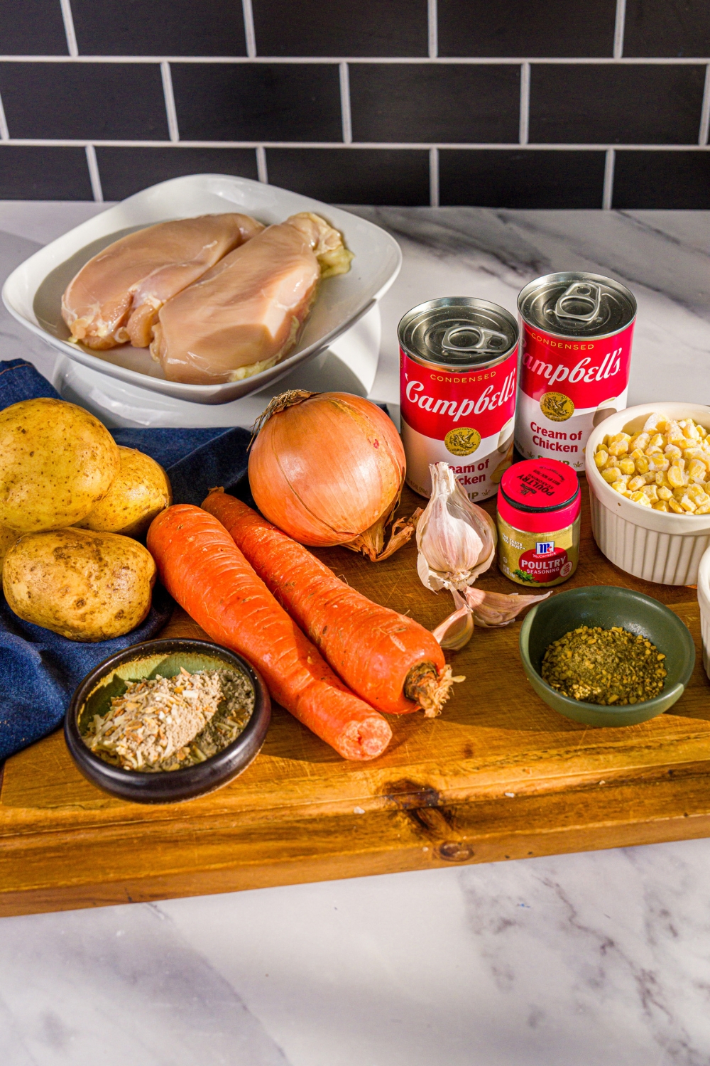 A wooden board with ingredients to make crockpot chicken pot pie including chicken breasts, cream of chicken soup, garlic, potatoes, frozen corn, onion, and seasonings.
