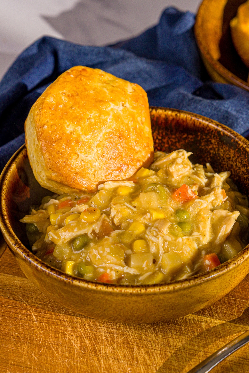A bowl of crockpot chicken pot pie served with a biscuit. The bowl is on a wooden board with a blue cloth napkin.