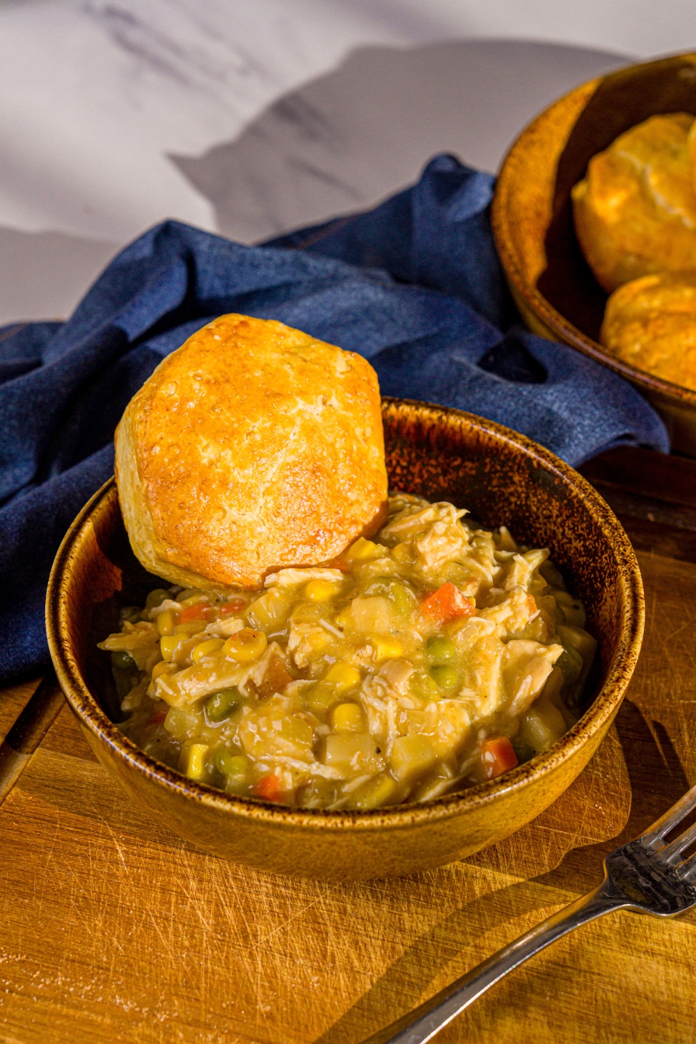 A bowl of crockpot chicken pot pie served with a biscuit. The bowl is on a wooden board with a fork and blue cloth napkin.