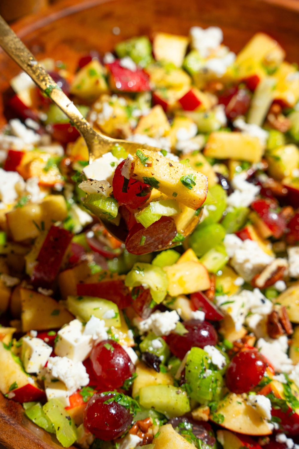 A close up of a spoon with a bite of apple crunch salad including apples, grapes, pecans, feta cheese, celery, and parsley in a maple dijon dressing. There is a bowl of salad in the background.