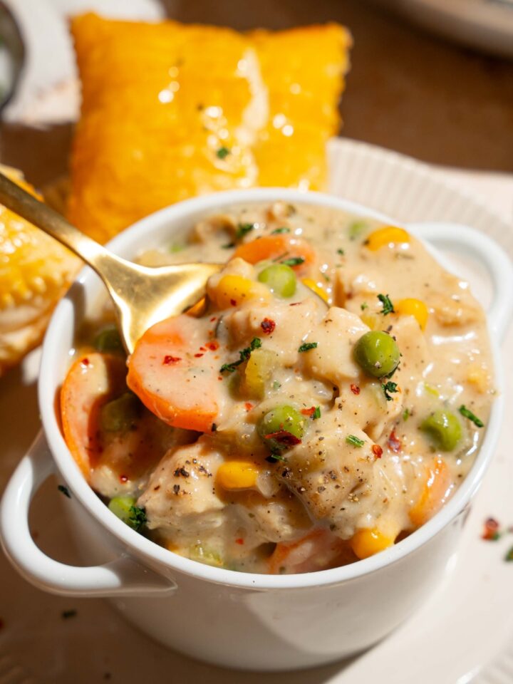 A small pie tin with crustless chicken pot pie garnished with pepper. A spoon is taking a bite from the tin. The tin is on a plate with a puff pastry.