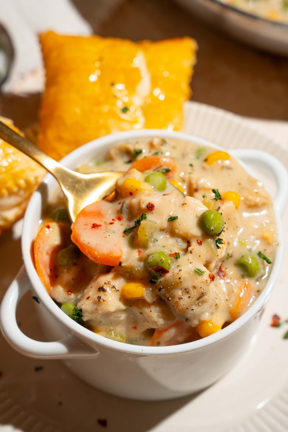 A small pie tin with crustless chicken pot pie garnished with pepper. A spoon is taking a bite from the tin. The tin is on a plate with a puff pastry.