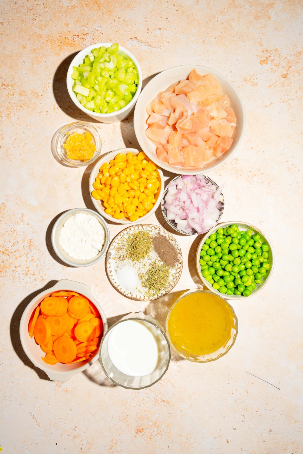 A tan counter with several bowls containing ingredients to make crustless chicken pot pie including chicken, peas, celery, onion, corn, broth, carrots, milk, and seasonings.