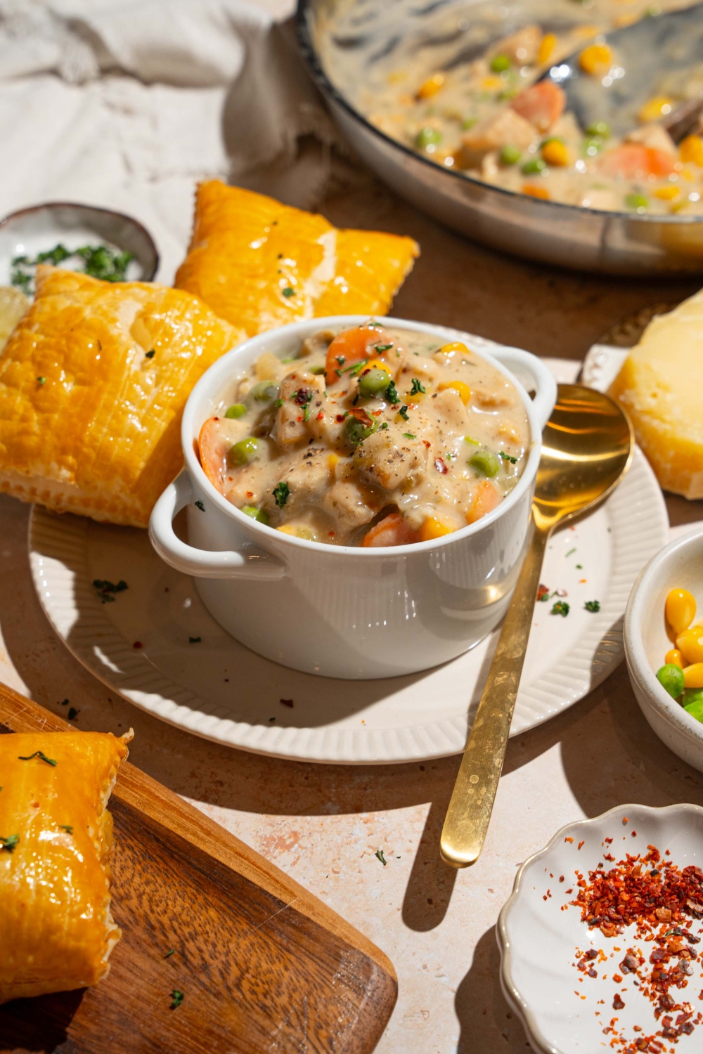 A small pie tin with crustless chicken pot pie garnished with pepper. The tin is on a plate with puff pastry and a spoon. The plate is on a tan counter with small plates of ingredients.