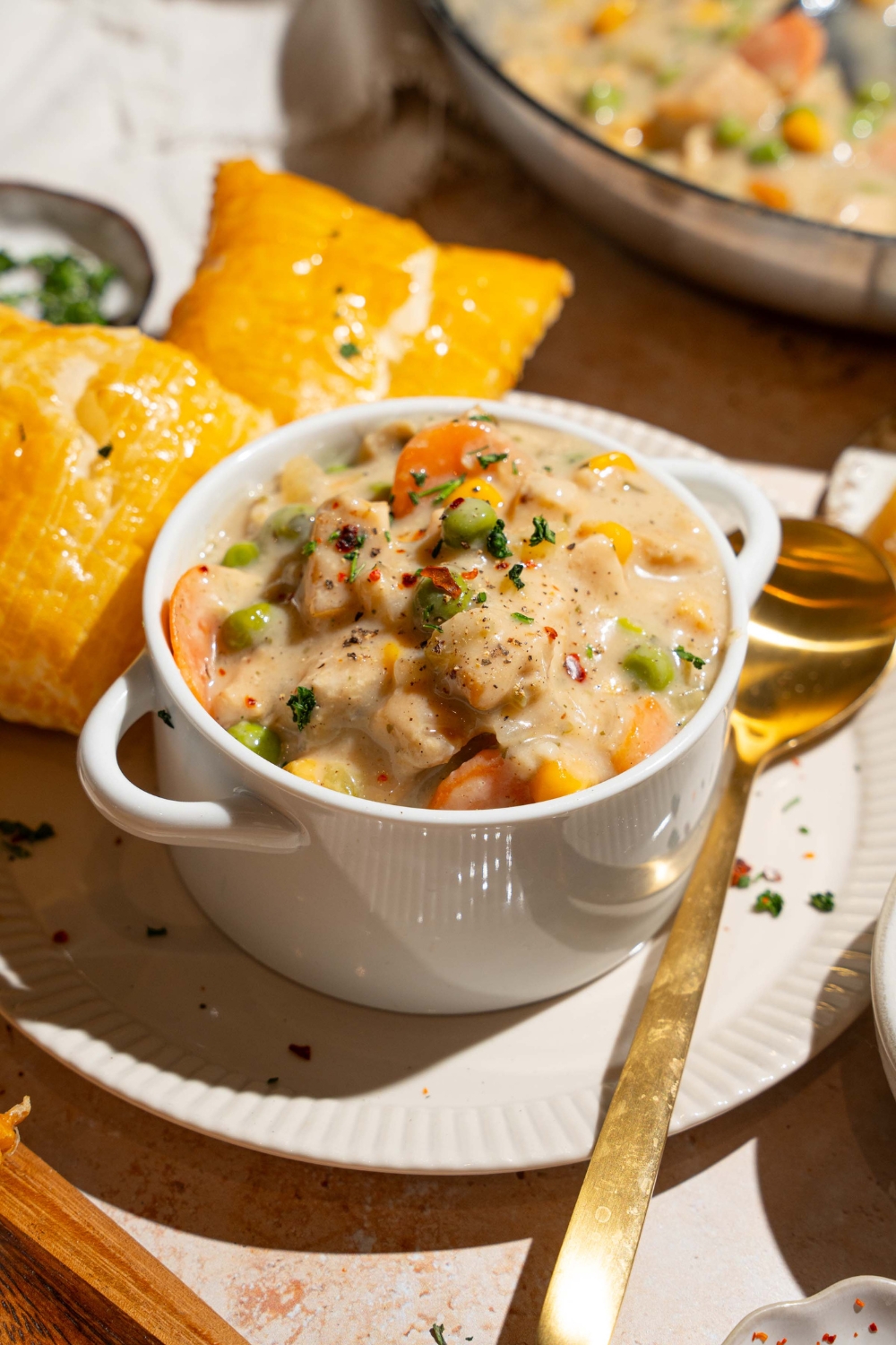 A small pie tin with crustless chicken pot pie garnished with pepper. The tin is on a plate with puff pastry and a spoon. The plate is on a tan counter with small plates of ingredients.