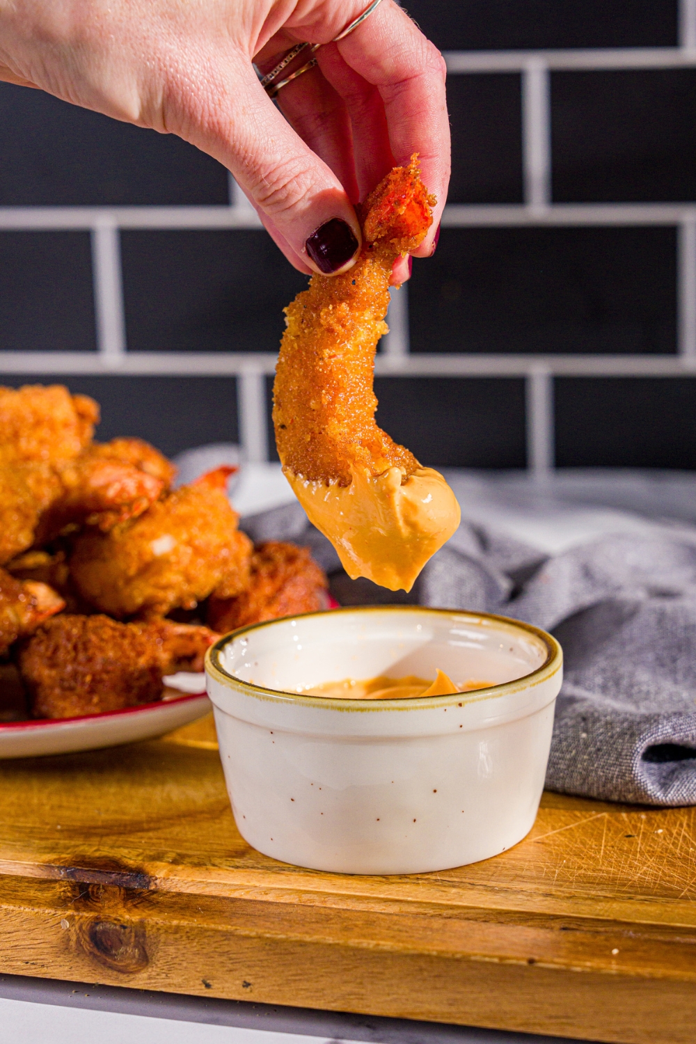 A hand dipping a crispy fried prawn into a bowl of spicy mayo. The bowl is on a wooden board with a plate of prawns and a blue cloth napkin.