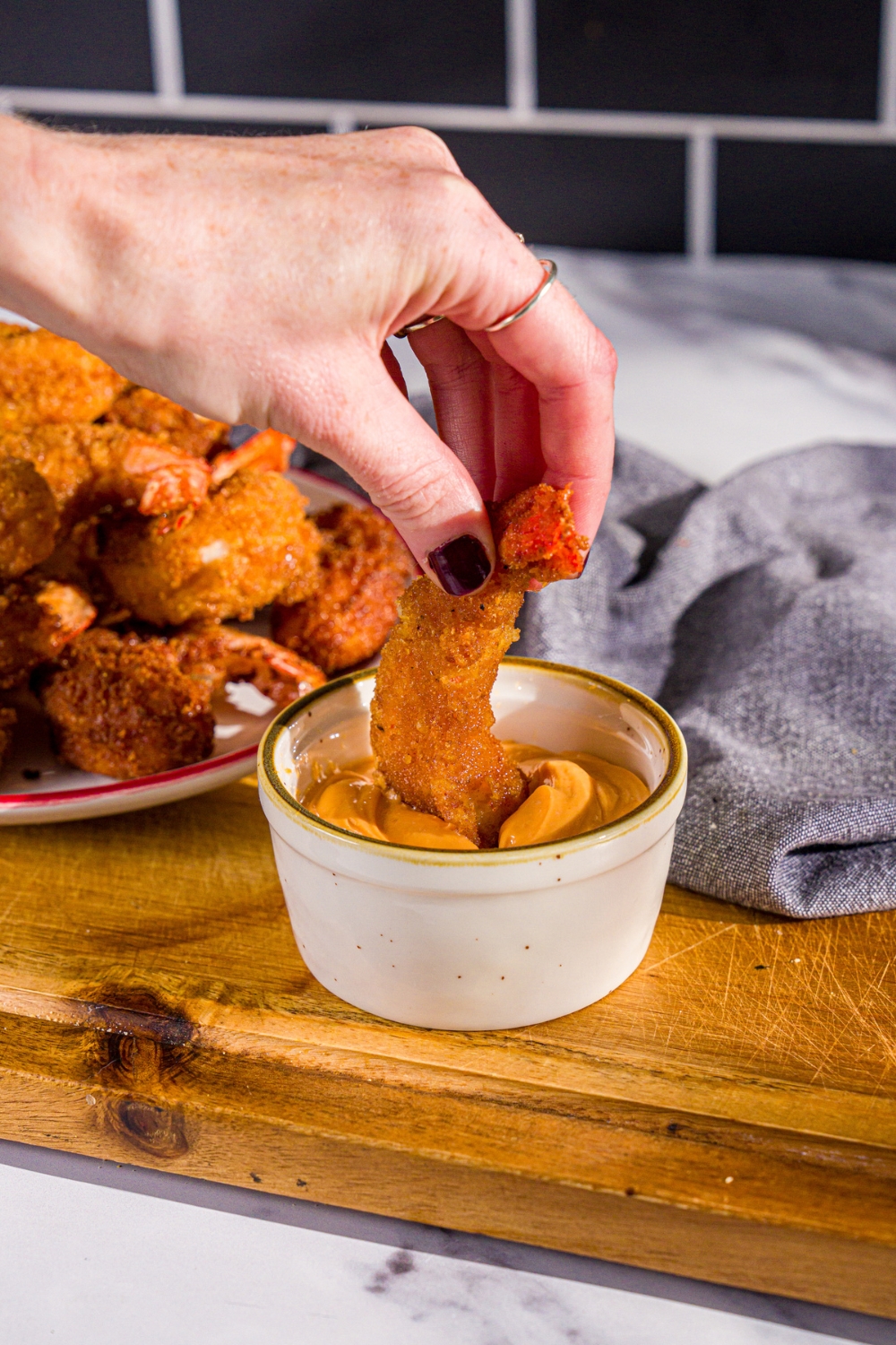 A hand dipping a crispy fried prawn into a bowl of spicy mayo. The bowl is on a wooden board with a plate of prawns and a blue cloth napkin.