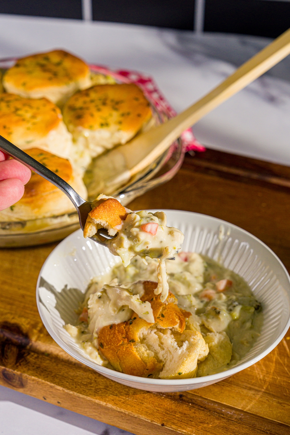A bowl of Trader Joe's chicken pot pie with a fork taking a bite from the bowl. The bowl is on a wooden board with a round baking dish with chicken pot pie topped with biscuits.