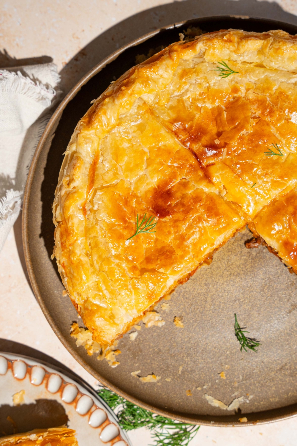 A pie plate with a whole beef pot pie garnished with fresh herbs. A slice of pie has been removed from the plate. The plate is on a tan counter with a white cloth napkin.