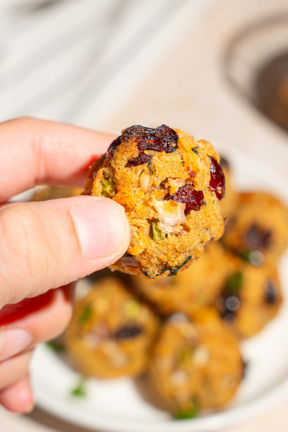 A close up of a hand holding a cranberry turkey stuffing ball. There is a plate of stuffing balls on a tan counter blurred in the background.