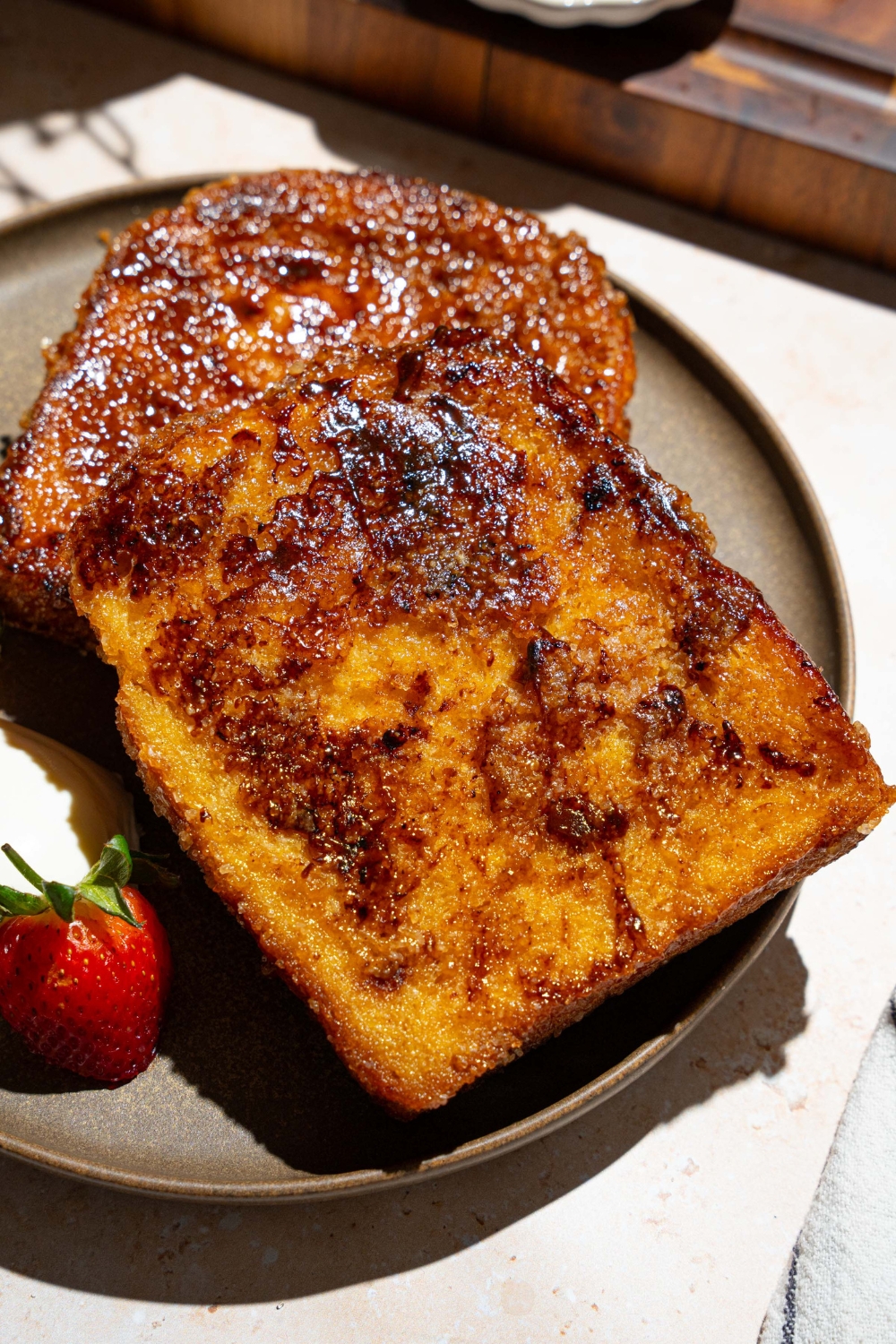 A plate with slices of creme brulee french toast served with sliced strawberries and whipped cream. The plate is on a tan counter.