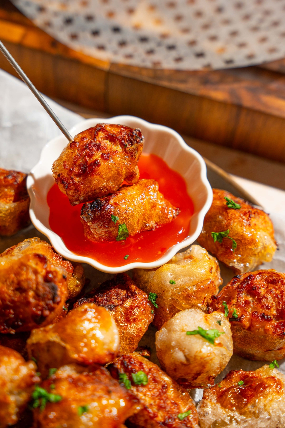 A skewer with crispy rice paper chicken bites dipping into a small bowl of dipping sauce. The bowl is on a plate of chicken bites.