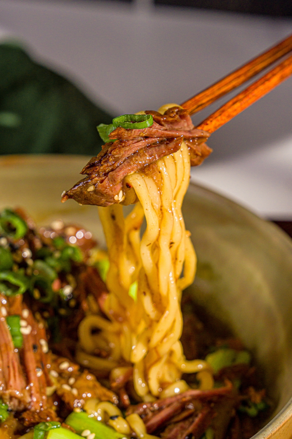 A bowl of crockpot Mongolian beef served with noodles and garnished with sliced green onions and sesame seeds. A pair of chopsticks are pulling a bite from the bowl.