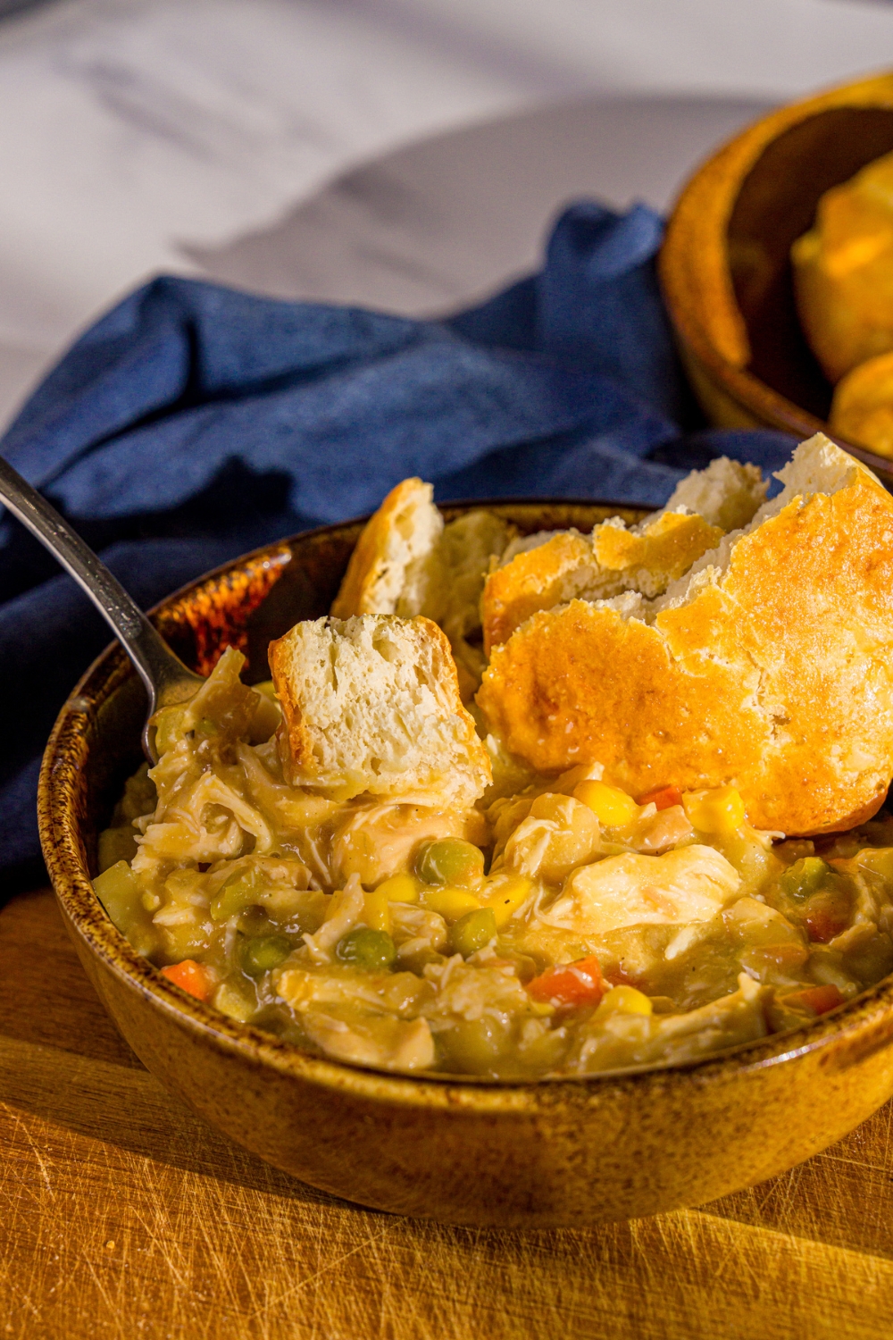 A bowl of crockpot chicken pot pie topped with a biscuit with a fork in the bowl. The bowl is on a wooden board with a blue cloth napkin.