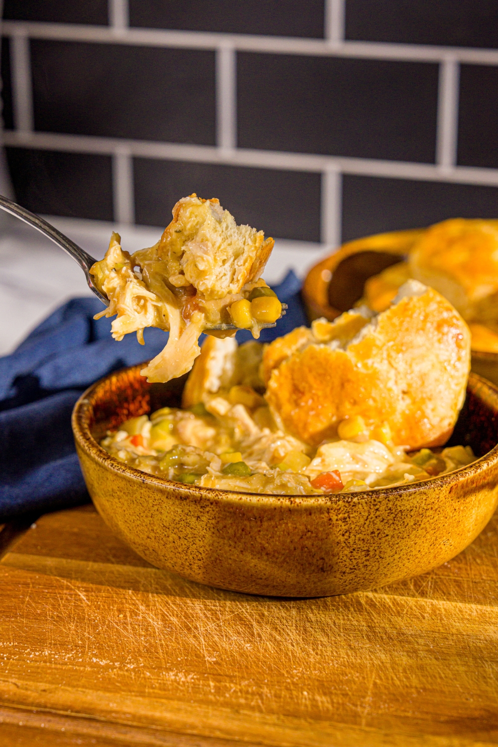 A spoon with a bite of crockpot chicken pot pie topped with a piece of biscuit. There is a bowl of pot pie on a wooden board with a blue cloth napkin.