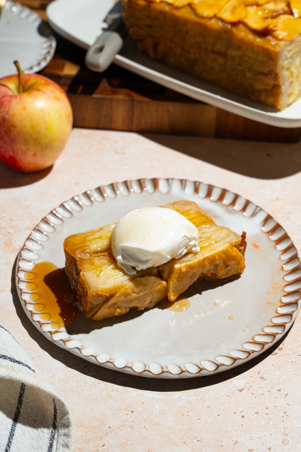 A white plate with a slice of invisible apple cake topped with vanilla ice cream. The plate is on a tan counter with a platter of cake and an apple.