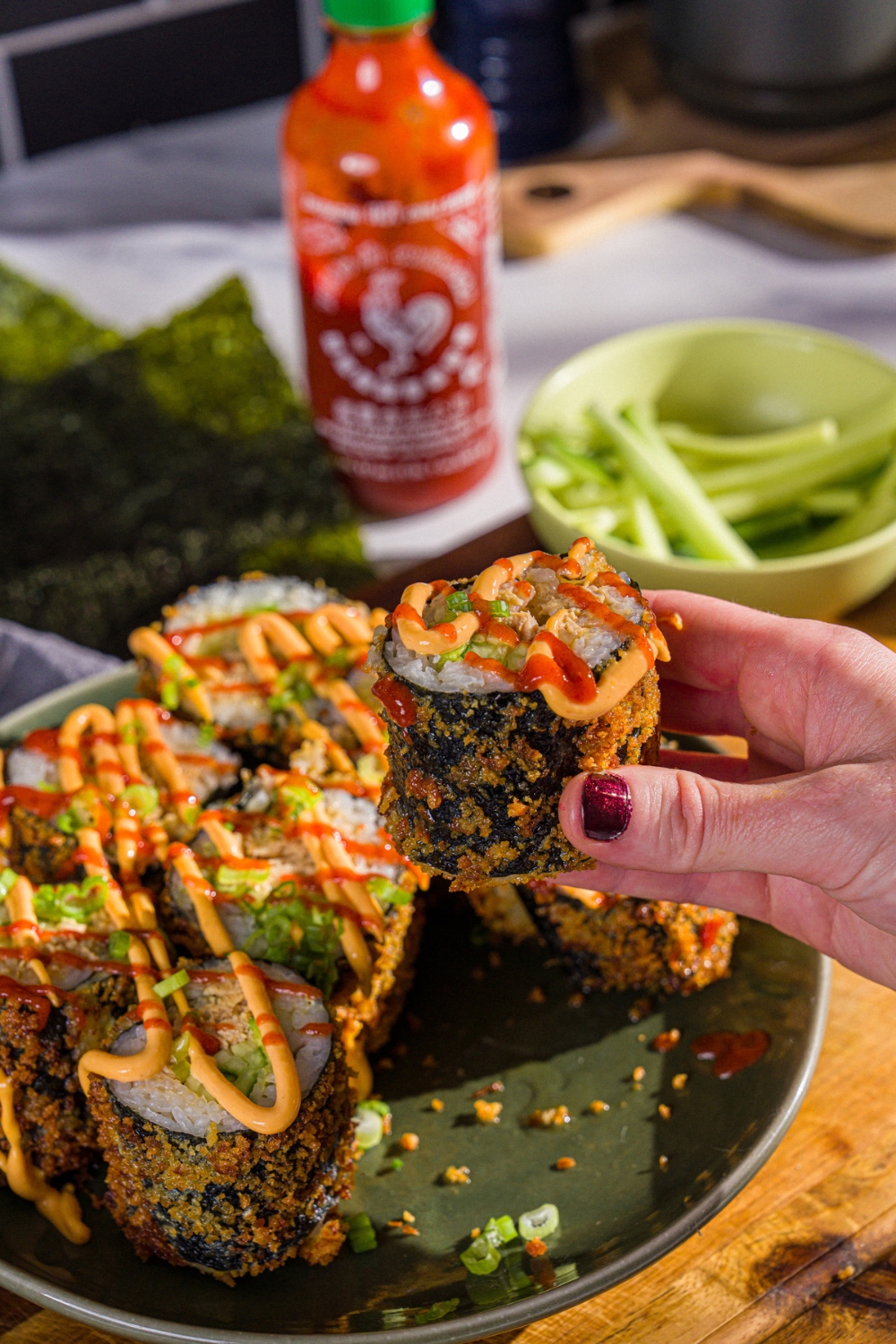 A hand taking a fried sushi burrito from a plate of sushi. The sushi is topped with sriracha, spicy mayo, and sliced green onion. The plate is on a wooden board.