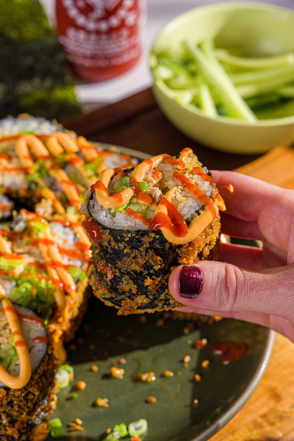 A hand taking a fried sushi burrito from a plate of sushi. The sushi is topped with sriracha, spicy mayo, and sliced green onion. The plate is on a wooden board.
