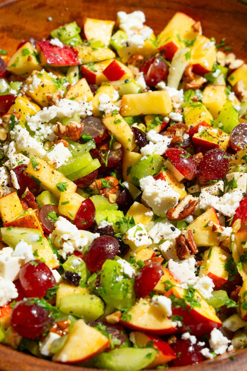 A wooden bowl with apple crunch salad including apples, celery, pecans, feta cheese, parsley, and grapes tossed in a maple dijon dressing. The bowl is on a tan counter with grapes, pecans, and lemon.