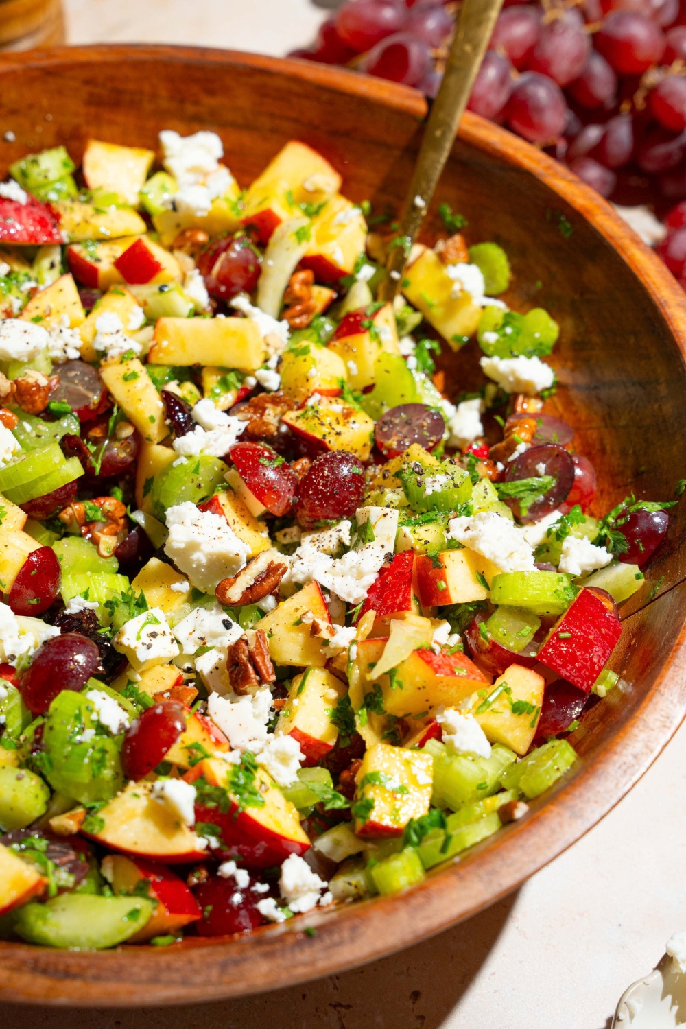 A wooden bowl with apple crunch salad including apples, celery, pecans, feta cheese, parsley, and grapes tossed in a maple dijon dressing. There is a fork in the bowl. The bowl is on a tan counter with grapes.