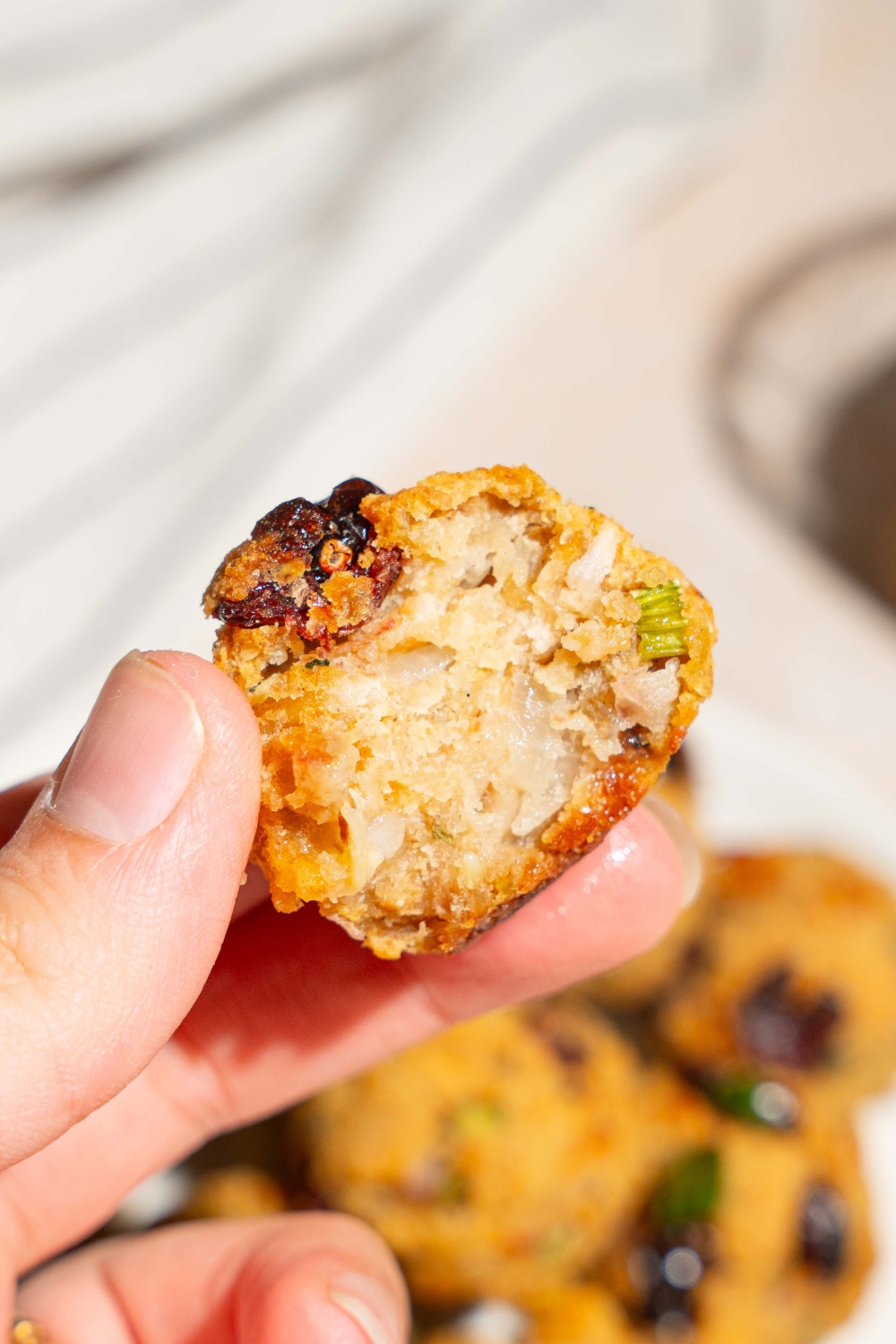 A close up of a hand holding a cranberry turkey stuffing ball with a bite removed. There is a plate of stuffing balls on a tan counter blurred in the background.