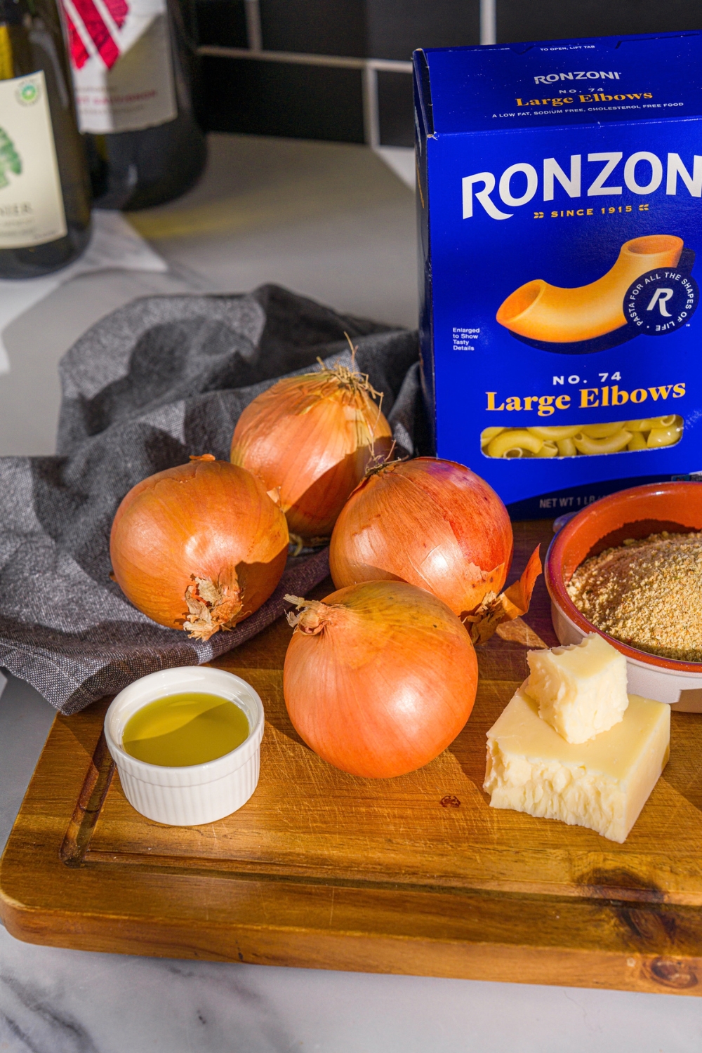 A wooden board with ingredients for French onion mac and cheese including onions, breadcrumbs, oil, cheese, and macaroni.