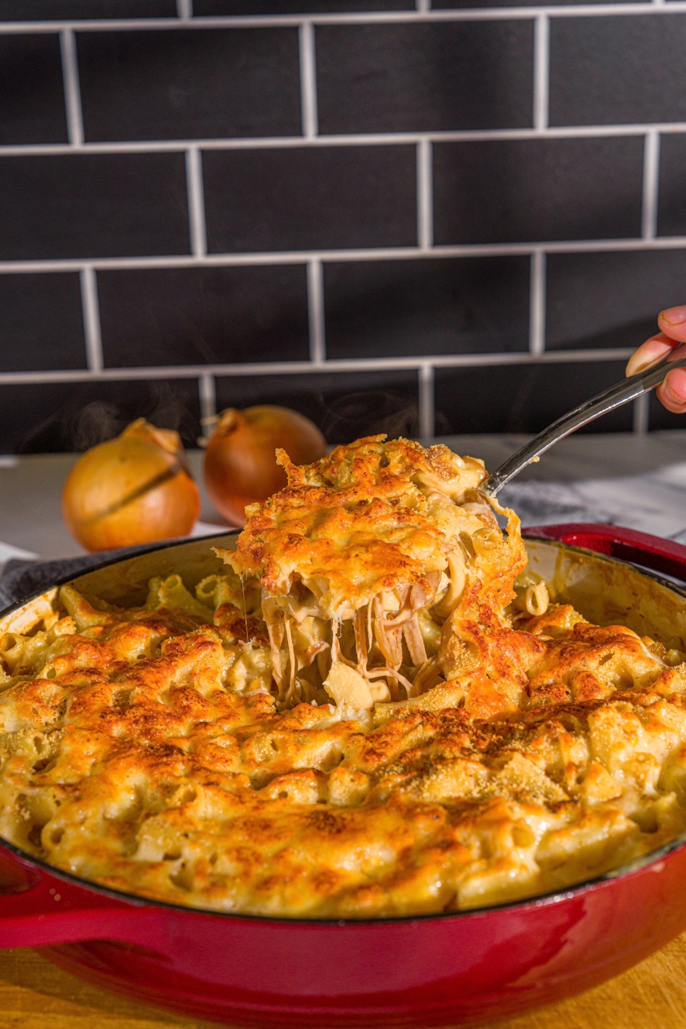 A cast iron pan with French onion mac and cheese. A spoon is taking a bite from the skillet. The skillet is on a wooden board.