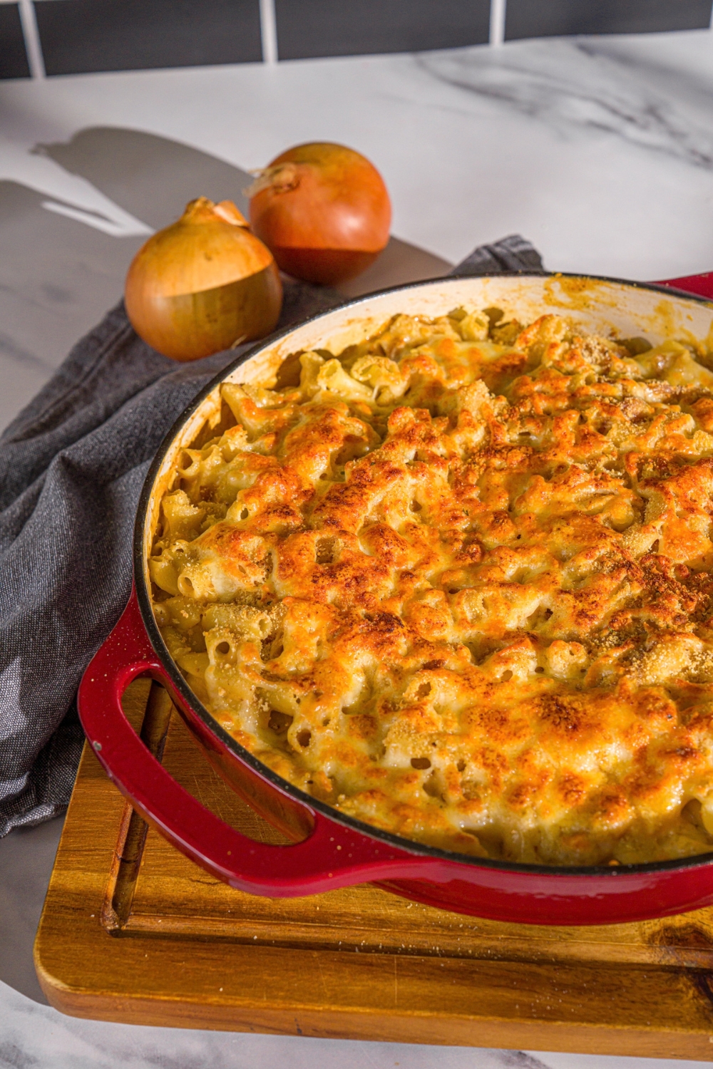 A cast iron pan with French onion mac and cheese. The skillet is on a wooden board with a blue cloth napkin and onions.