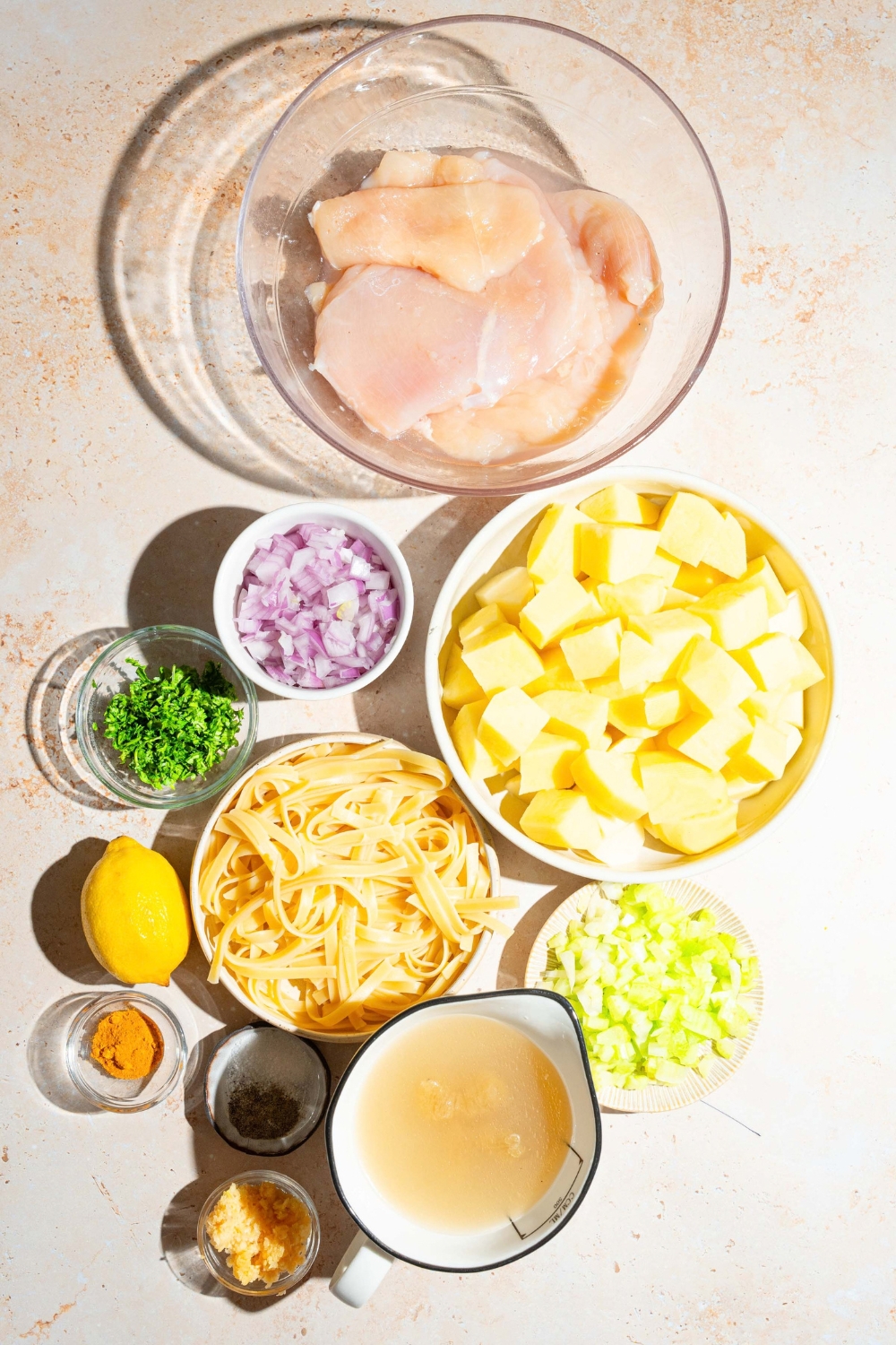 A tan counter with ingredients to make garlic chicken soup including chicken, potatoes, garlic, chicken broth, onion, lemon, celery, noodles, and more.