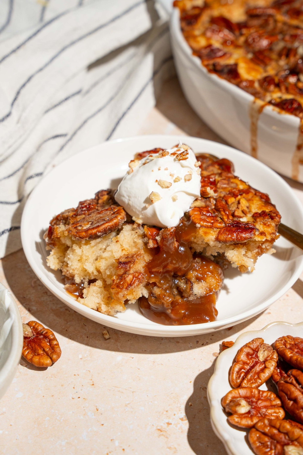 A white plate with a slice of pecan pie cobbler topped with a dollop of whipped cream with chopped pecans. There is a spoon on the plate. The plate is on a tan counter with a casserole dish of cobbler and a white striped napkin.