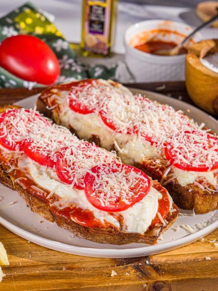A plate with two slices of Hailey Bieber pizza toast finished with shredded parmesan. The plate is on a wooden board with a bowl of salt, marinara sauce, and a tomato.