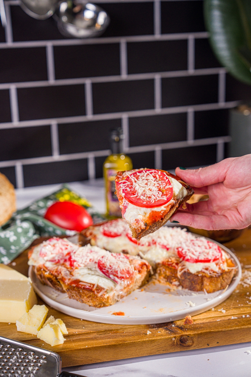 A close up of a hand holding a slice of Hailey Bieber pizza toast topped with shredded parmesan. There is a plate of pizza toast on a wooden board blurred in the background.