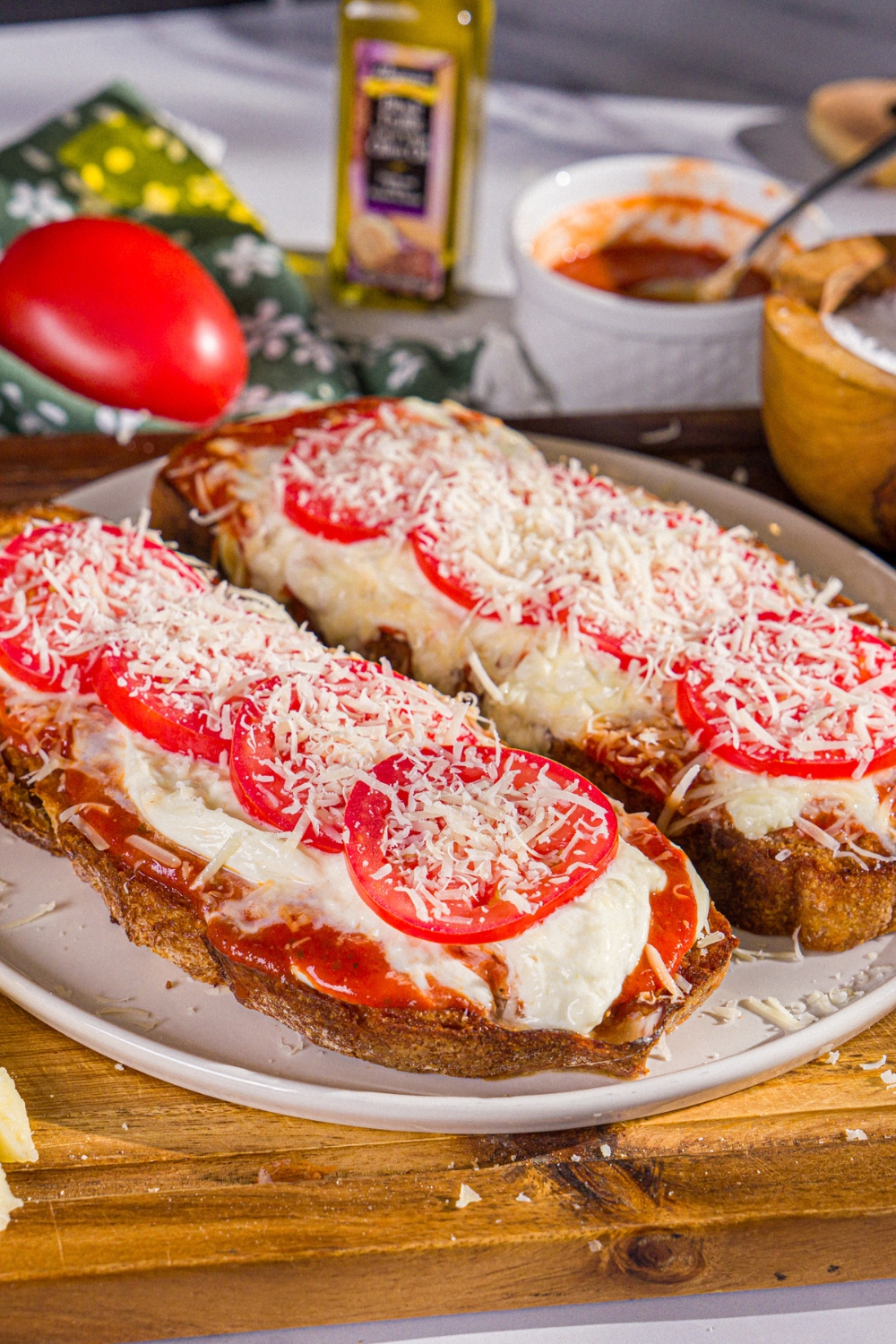 A plate with two slices of Hailey Bieber pizza toast finished with shredded parmesan. The plate is on a wooden board with a bowl of salt, marinara sauce, and a tomato.