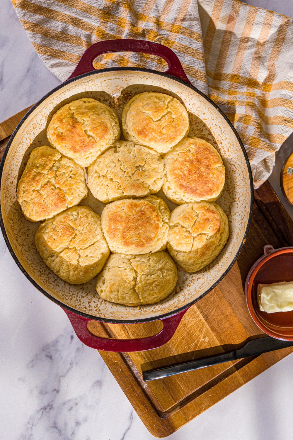 A baking dish with baked Hardee's biscuits. The dish is on a wooden board with a yellow checkered napkin and small bowl of butter.