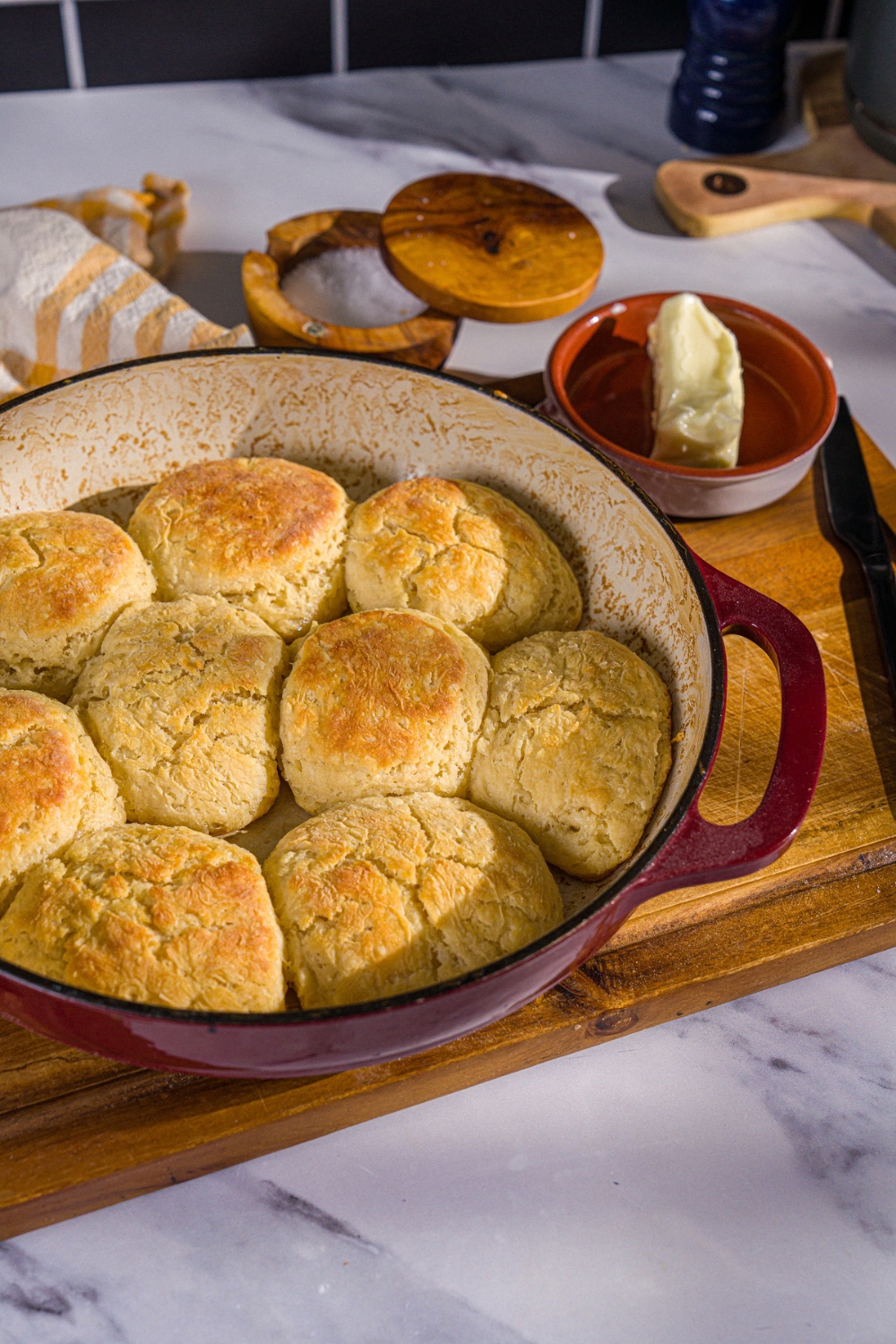 A baking dish with baked Hardee's biscuits. The dish is on a wooden board with a yellow checkered napkin and small bowl of butter.