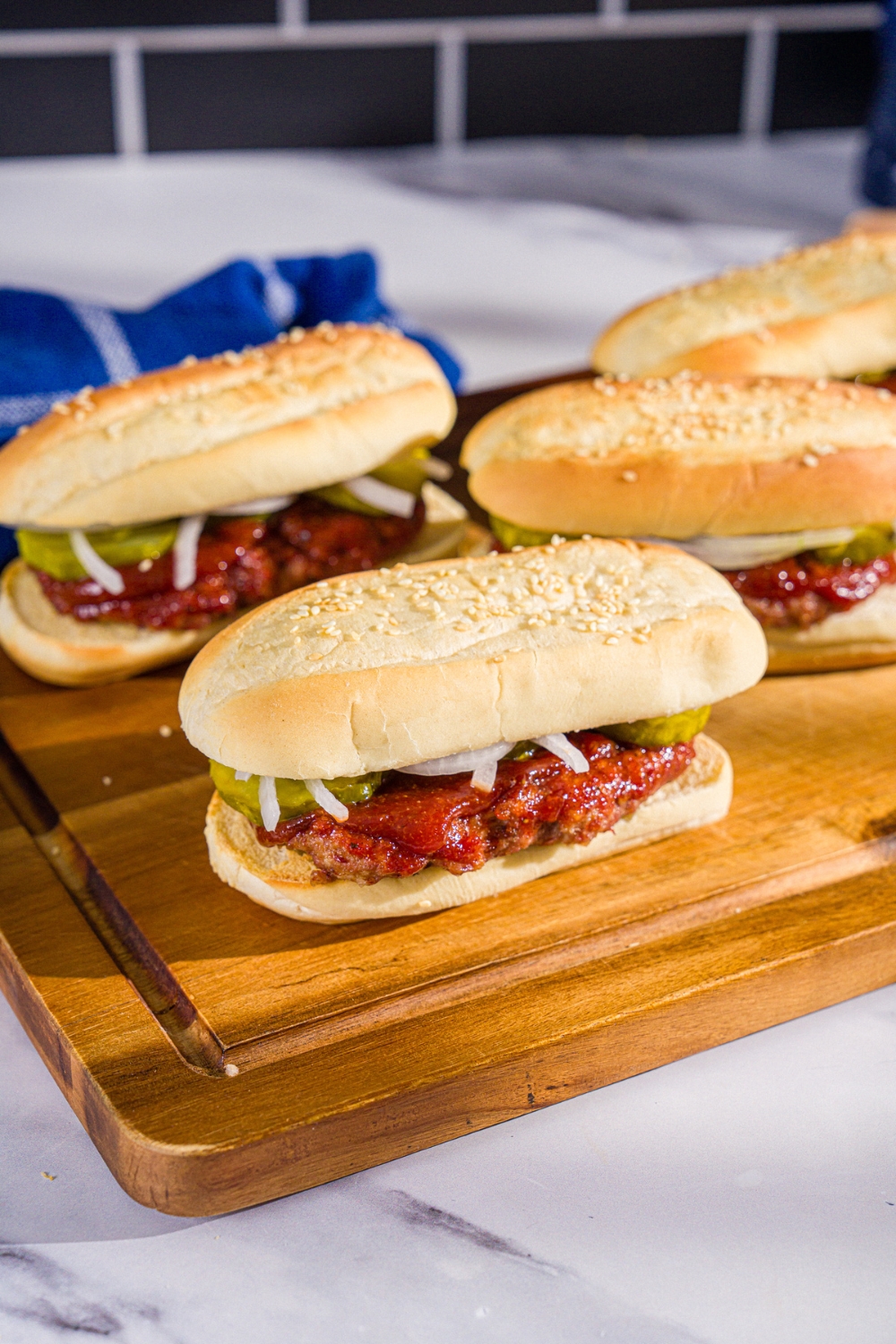 A wooden board with several homemade McRib sandwiches on a hoagie roll with pickles, onions, and homemade BBQ sauce. The board is on a marble counter with a blue cloth napkin.