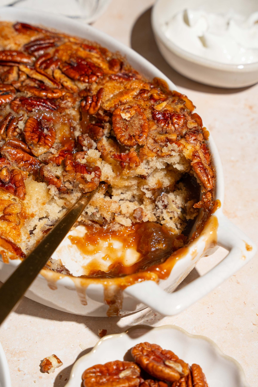A casserole dish with pecan pie cobbler with a section removed. A serving spoon is dipped in the cobbler. The dish is on a tan counter with a bowl of pecans and bowl of whipped cream.