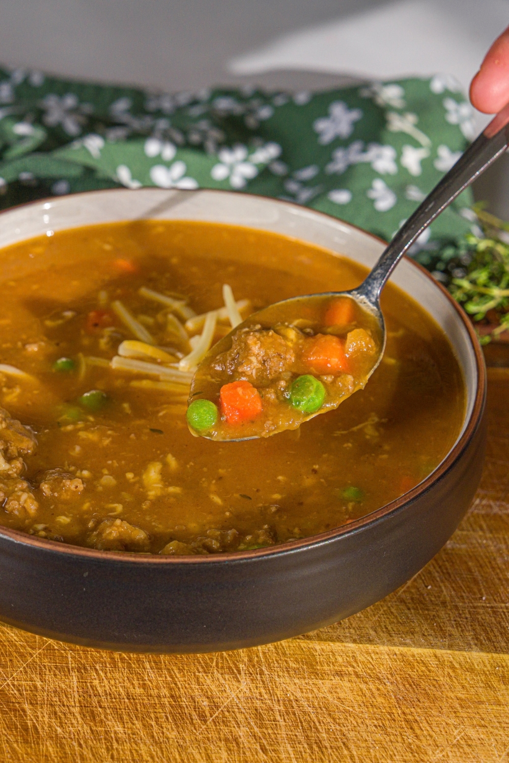 A bowl of shepherd's pie soup topped with shredded cheese. A spoon is taking a bite from the bowl. The bowl is on a wooden board with a green cloth napkin.