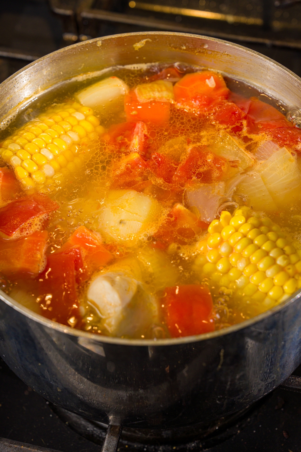 A stock pot with ABC soup with corn on the cob, tomatoes, potatoes, chicken, and carrots simmering on the stove.