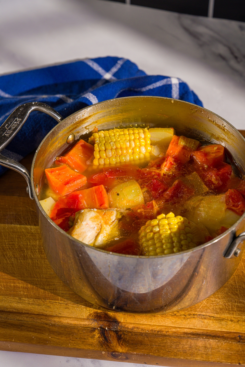 A pot of ABC soup with corn on the cob, carrots, potatoes, and chicken. The pot is on a wooden board with a blue cloth napkin.
